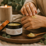 A person applies a pale yellow-green salve from a jar labeled 'Cannabis Craft' onto their hand, set on a wooden tray with a candle, spoon, and eucalyptus leaves.
